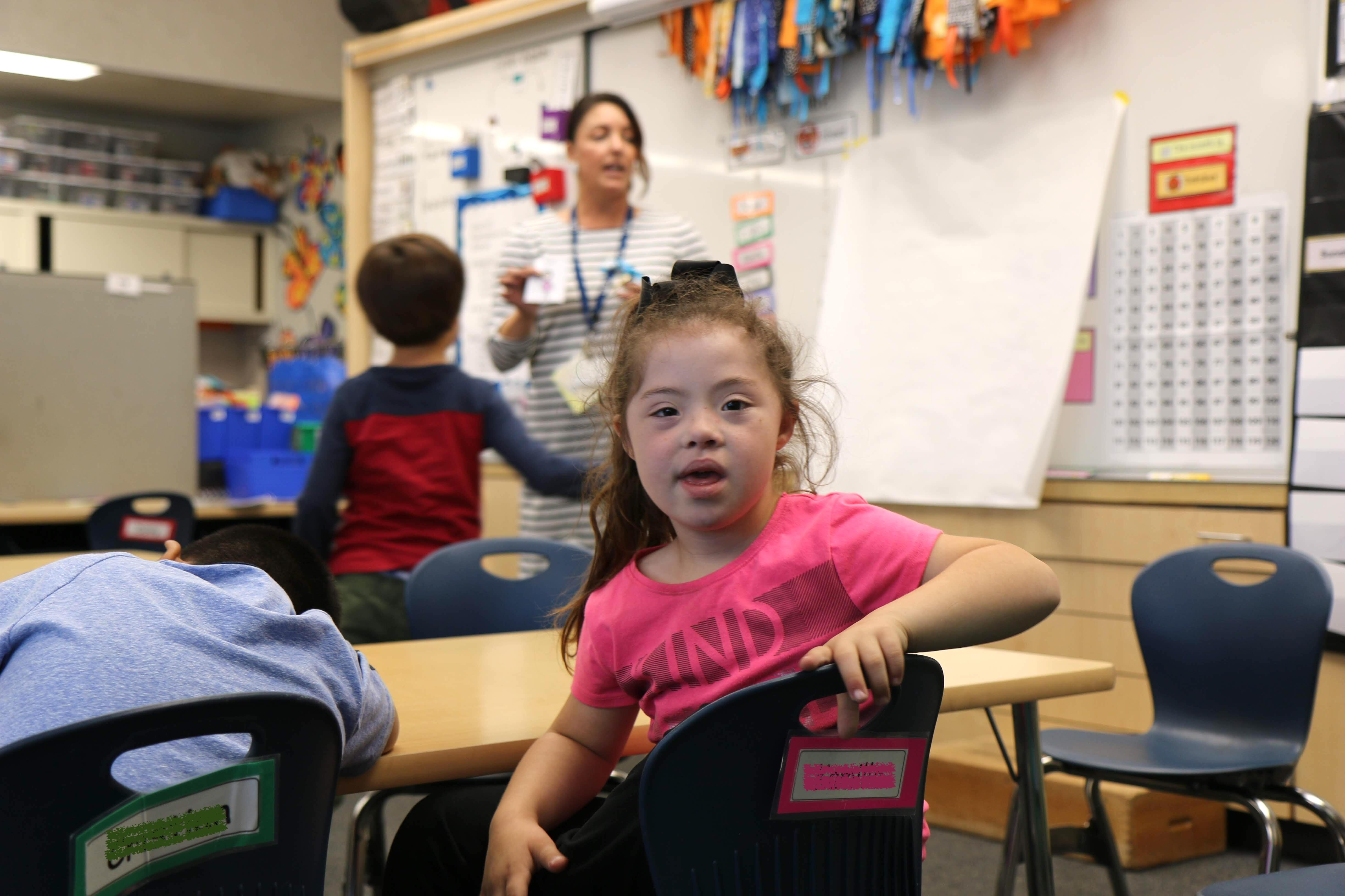 little girl with Down Syndrome in classroom looks at the camera as her teacher talks in the background.