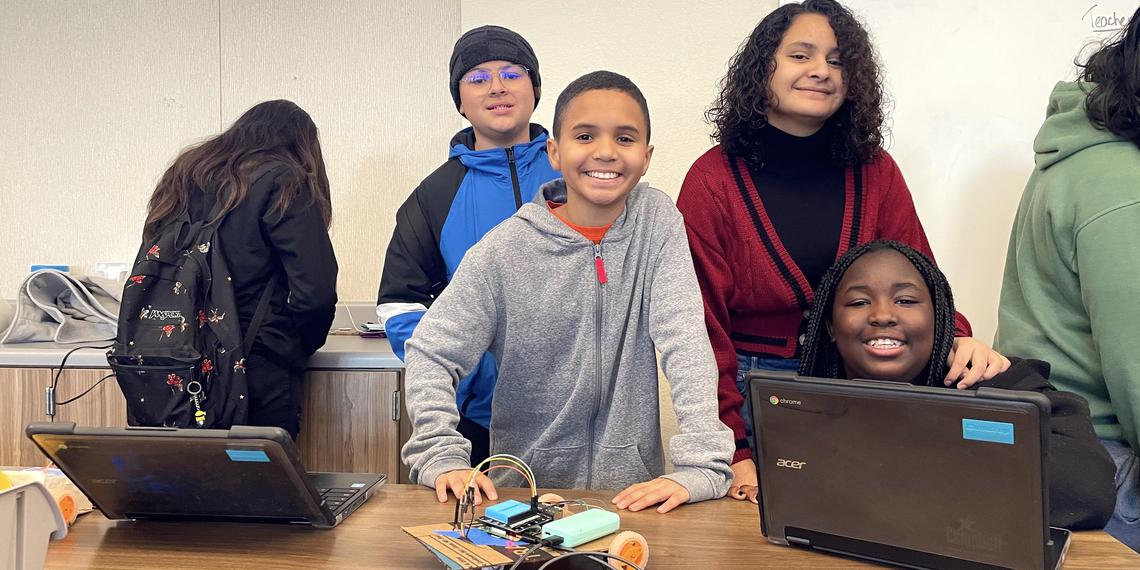 four middle school students smile beside their laptop and hand-made and coded robot vehicle