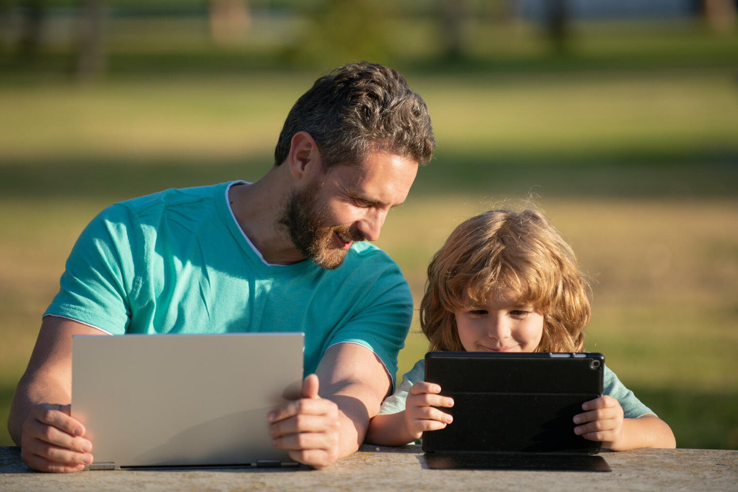 father and son smiling with laptops in the park